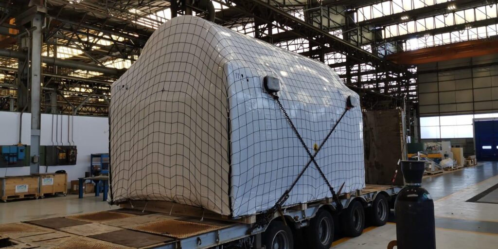 Large industrial warehouse with a massive wrapped cargo secured on a multi‑axle trailer using a net and chains, with a tall gas cylinder in the foreground.