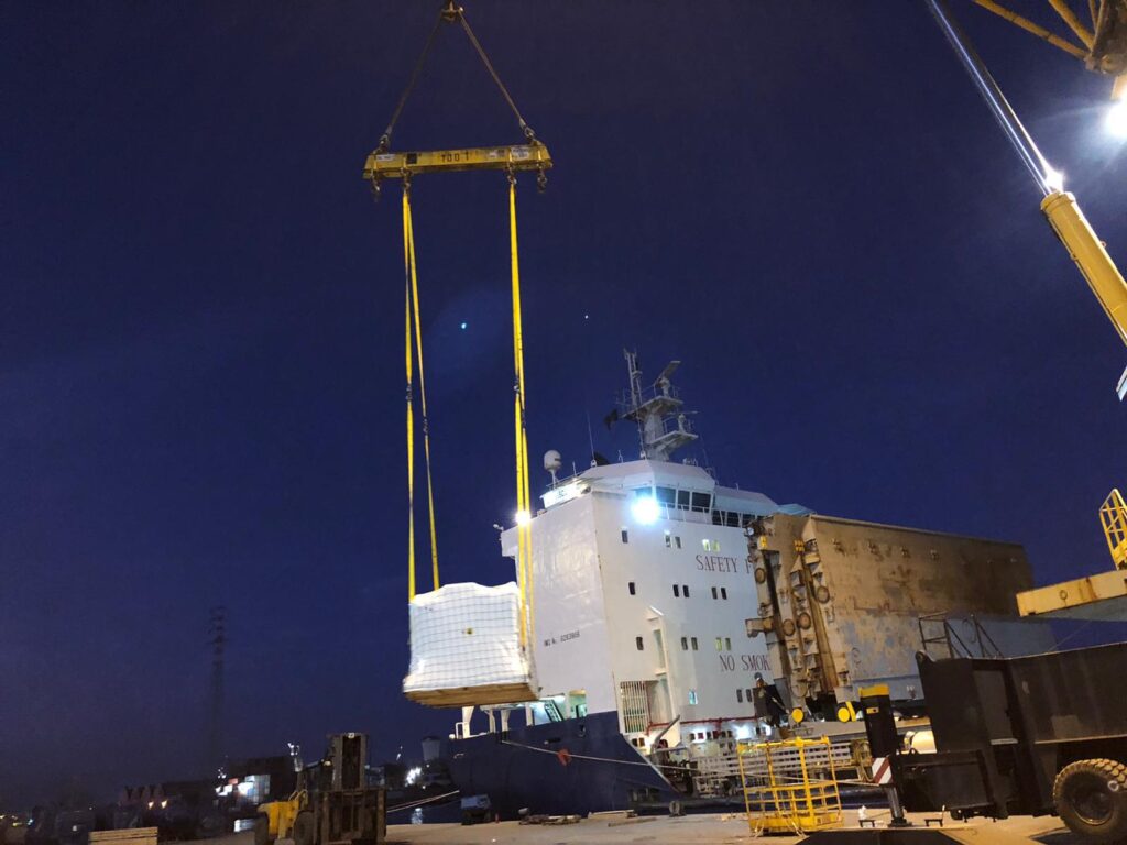 Nighttime shipyard scene with a large crane lifting a yellow frame over a vessel labeled SAFETY; dark sky, port equipment nearby.