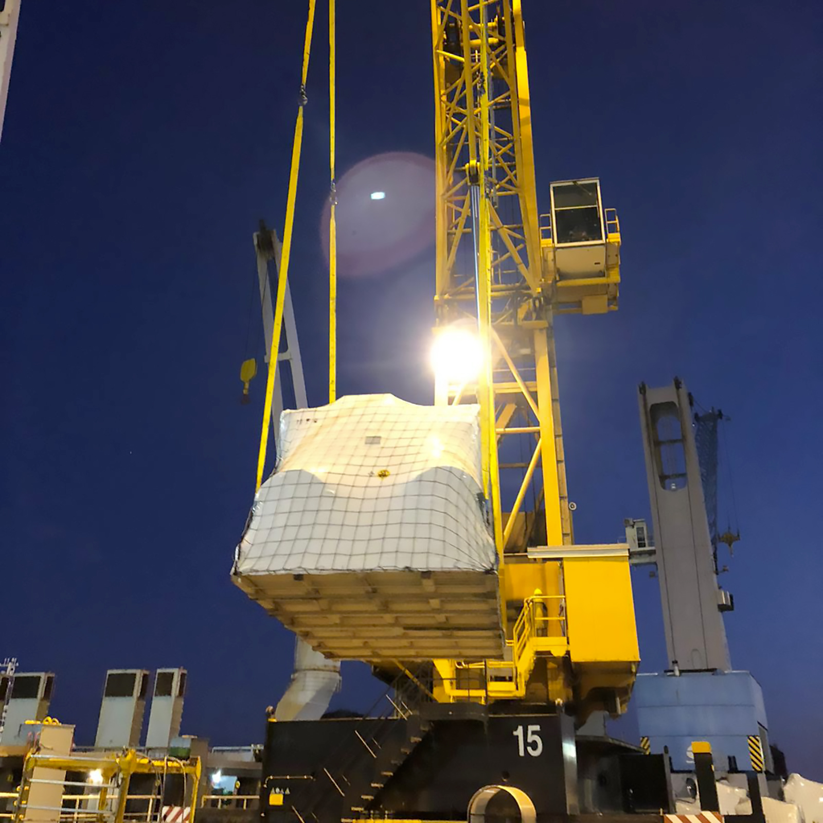 Nighttime construction crane with a yellow lattice boom lifting a white netted load against a dark blue sky.